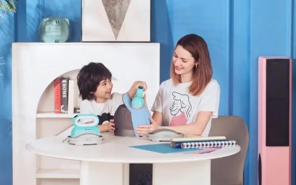 Woman and child in a homely setting. The child is pressing the LOKLiK ImPress™ Hat on a light blue hat. A second LOKLiK ImPress™ Hat is placed on the table and sits on its safety base.