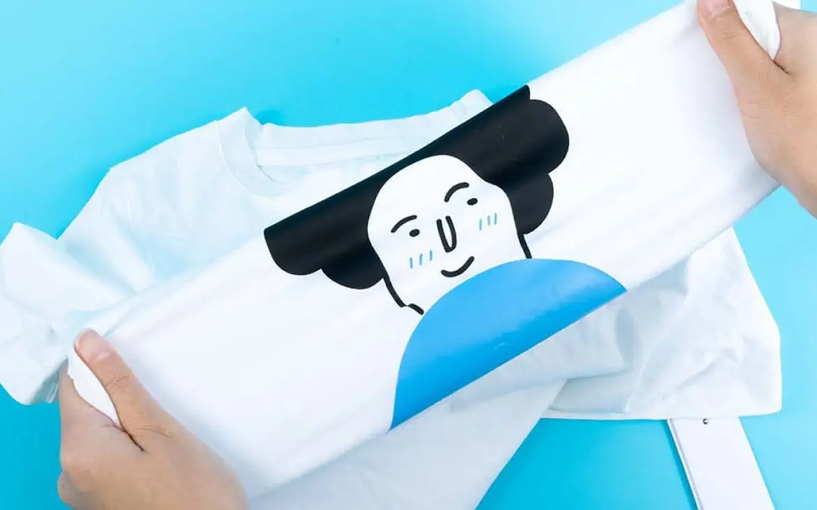 Two hands stretching the fabric of a white shirt with an heat transfer vinyl design of an illustrated face.