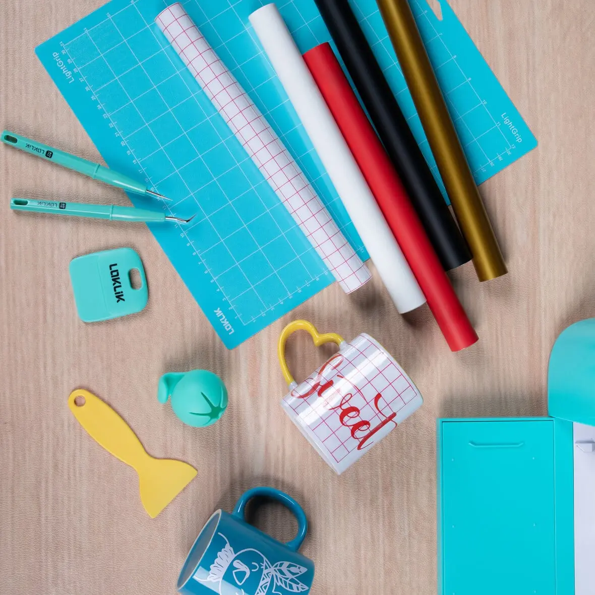 Top down view of a wooden table with a LOKLiK Crafter in LOKLiK Blue with a green cutting mat. There are 4 rolls of vinyl, a roll of grid transfer tape, weeding tools, scrap collector and a white mug with grid transfer tape applied.