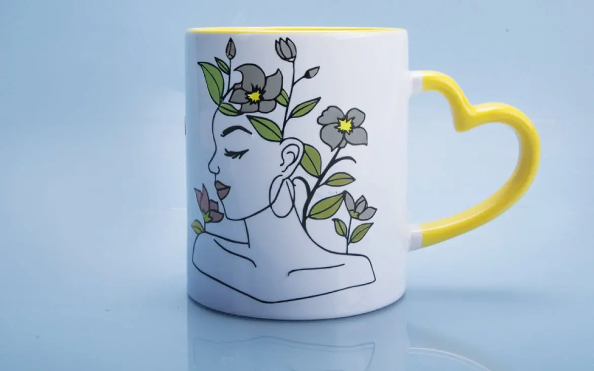 Frontal view of a white mug with a yellow heart shaped handle with a desingn in glossy permanent adhesive vinyl of a woman's face with flowers as hair.