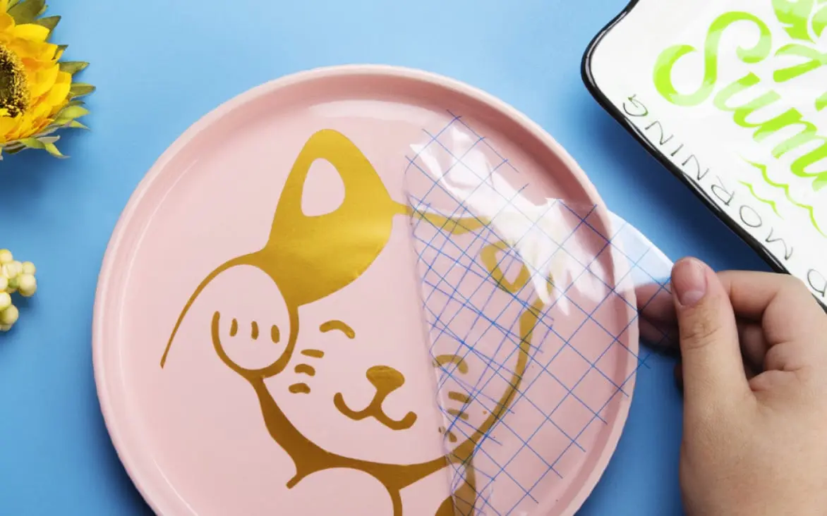 Top view of a pink plat on a blue background. A hand is peeling of grid transfer tape of a design of a lucky cat.