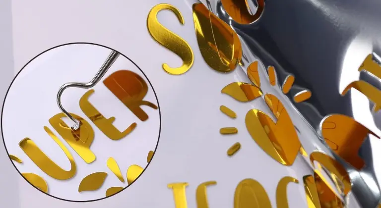Close-up of a weeding hook pulling a piece of superfluous vinyl from the center of the letter P made from gold mirror metallic permanent adhesive vinyl.